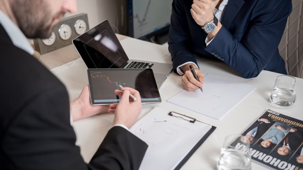 Two businessmen discussing financial charts on a tablet in a modern office setting.